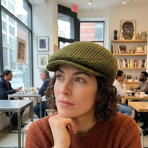 May include: A woman wearing a green, textured newsboy cap and a rust-colored sweater. She is sitting in a cafe, with other people visible in the background. The cap has a rounded crown and a small, stiff brim.