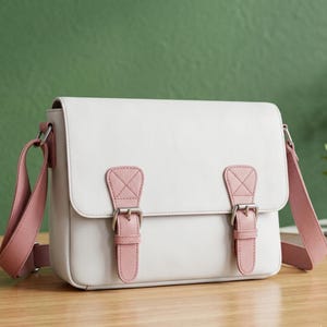 May include: A white satchel bag with pink accents. The bag features a flap closure with two pink straps and silver buckles. The shoulder strap is also pink. The bag is made of a smooth material and has a rectangular shape.