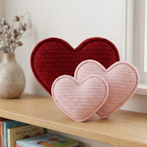 May include: Three heart-shaped crochet pillows in varying sizes and shades of red and pink. The largest pillow is a deep red, while the two smaller ones are light pink. The pillows are displayed on a wooden shelf.
