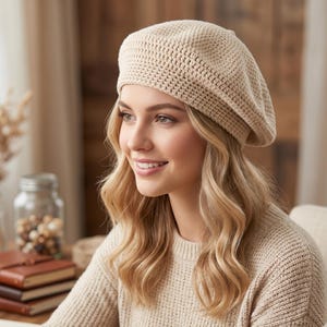 Op de afbeelding: Een beige gehaakte baret. De baret wordt gedragen door een persoon met blond haar en een bijpassende beige trui. De hoed heeft een getextureerd uiterlijk en is gemaakt van een zacht, gebreid materiaal. De afbeelding is binnenshuis gemaakt.
