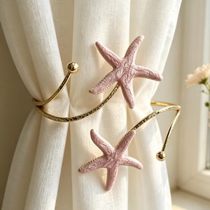 May include: Gold-coloured metal curtain tiebacks featuring pink starfish. The tiebacks have a curved design with decorative detailing and small gold spheres. The starfish are a muted pink colour, adding a coastal touch.
