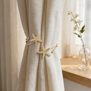 Può includere: Due fermatende dorati a forma di stella marina. I fermatende tengono indietro una tenda color crema. Un piccolo vaso di vetro con fiori bianchi è appoggiato su un davanzale di legno sullo sfondo.