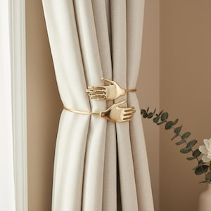 Puede incluir: Sujetacortinas dorados con forma de manos que sujetan una cortina beige. Las manos, con dedos detallados, están unidas a una barra curva. La cortina cuelga frente a una pared de color neutro.