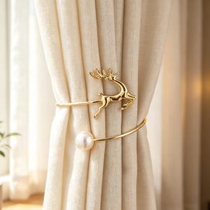 May include: Gold-colored curtain tiebacks with a deer silhouette and a pearl accent. The tiebacks are wrapped around a cream-colored curtain, adding a decorative touch to the window treatment.