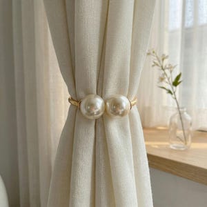 May include: Cream-coloured curtain tiebacks featuring large, faux pearl embellishments. The tiebacks are connected by a gold-toned metal band, offering a decorative and elegant appearance. The curtains are a light cream colour.