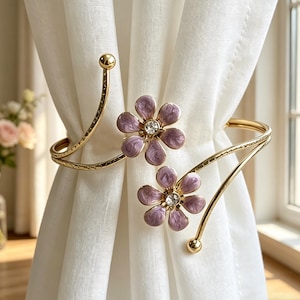 May include: Gold-toned metal curtain tiebacks with a floral design. Each tieback features a curved arm with a decorative flower in shades of lavender and a central crystal. The tiebacks are holding white curtains.