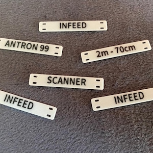 Puede incluir: Un conjunto de pequeñas etiquetas rectangulares blancas con texto negro. Las etiquetas incluyen las palabras "INFEED", "ANTRON 99", "SCANNER" y "2m - 70cm". Cada etiqueta tiene dos pequeños agujeros en cada extremo.