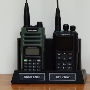 May include: Two-way radios, a green Baofeng and a black AnyTone, are in a black charging base. The Baofeng has a digital display and the AnyTone has a keypad. The base has the brand names printed on it.