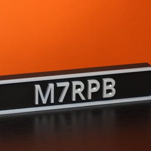 Puede incluir: Un letrero rectangular negro con las letras "M7RPB" en blanco. El letrero tiene un borde blanco y está sobre un fondo naranja. El letrero está sobre una superficie oscura.