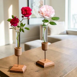 May include: Two glass test tube vases held by light wood stands with thin metal rods. One vase contains two vibrant red roses with green leaves, while the other holds a single pale pink rose with green leaves, creating a floral display.
