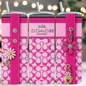 May include: A pink and white Coach tumbler with a floral design. The tumbler features a pink strap with silver buttons and a daisy charm. The Coach logo is displayed in silver. The tumbler has a silver straw.