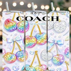 Könnte beinhalten: Ein weißer Becher mit einem sich wiederholenden Muster aus funkelnden, regenbogenfarbenen Kirschen und Kristallverzierungen. Das Design umfasst eine hellblaue Schleife und den Text "COACH NEW YORK". Der Becher hat einen silbernen Strohhalm.