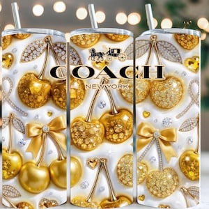 May include: A stainless steel tumbler with a decorative design featuring golden cherries, bows, and Coach New York branding. The design includes rhinestones and a white background, with a silver straw.