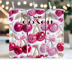 Puede incluir: Tres vasos de acero inoxidable con un diseño decorativo con cerezas, lazos y corazones. El diseño incluye el texto "COACH NEW YORK" y está adornado con pedrería y detalles dorados. Cada vaso tiene una pajita plateada.