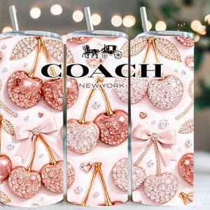May include: A set of three stainless steel tumblers with a decorative design. The design features sparkling pink cherries, bows, and the "COACH NEW YORK" logo. Each tumbler has a silver straw.