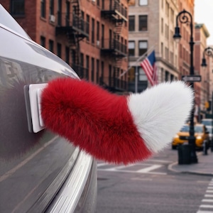 Puede incluir: Un accesorio de cola peluda roja y blanca sujeto a un coche. La cola es roja en la base y pasa a blanca en la punta. El coche es gris, y el fondo muestra una calle de la ciudad con edificios y una bandera.