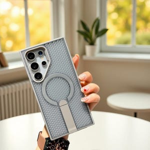 May include: A gray phone case with a textured surface and a circular ring holder. The case has a built-in stand and a camera cutout. The case is held in a person's hand.