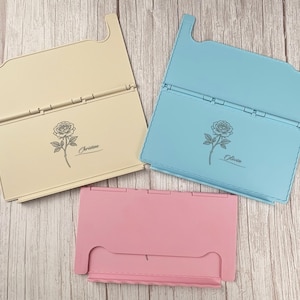 May include: Three rectangular folding stands in cream, light blue, and pink. The cream stand features a rose illustration and the name Christine, while the light blue stand features a rose illustration and the name Olivia. Each stand has a hinged design.