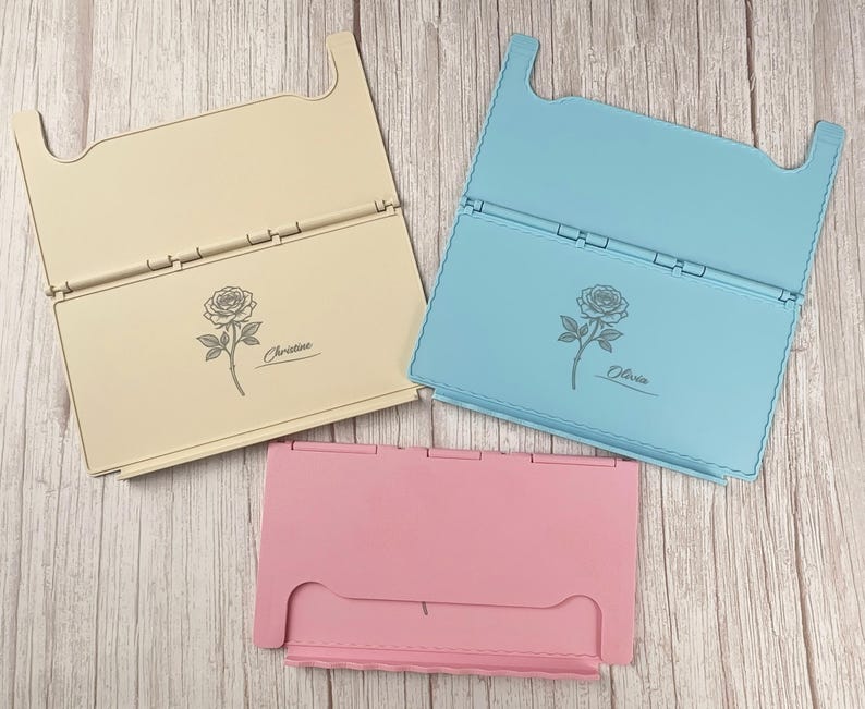 May include: Three rectangular folding stands in cream, light blue, and pink. The cream stand features a rose illustration and the name Christine, while the light blue stand features a rose illustration and the name Olivia. Each stand has a hinged design.