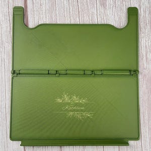 May include: An olive green foldable book stand featuring a hinged design and a horizontal ledge. The front panel is engraved with a botanical leaf illustration and the name Kathleen in a cursive script font, centred on the flat surface.