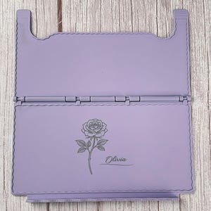 May include: A light purple, rectangular, hinged folding stand with a scalloped edge design. The bottom panel features a dark grey engraved illustration of a single rose with a stem and leaves, accompanied by the name Olivia in script text.