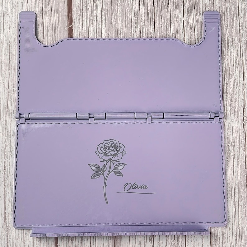 May include: A light purple, rectangular, hinged folding stand with a scalloped edge design. The bottom panel features a dark grey engraved illustration of a single rose with a stem and leaves, accompanied by the name Olivia in script text.