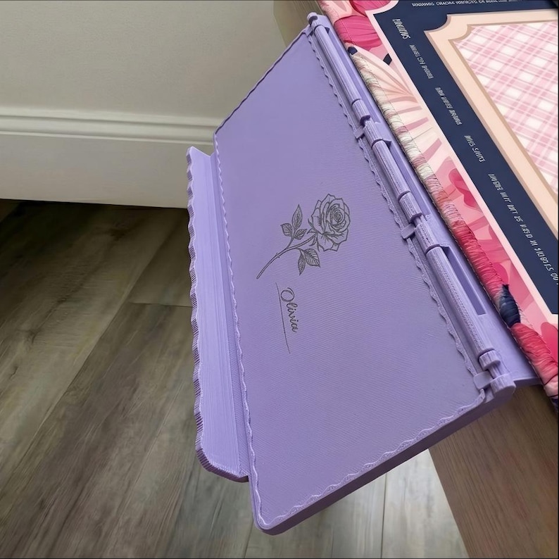May include: A light purple adjustable book stand featuring a laser-engraved rose illustration and the name Olivia in cursive script. The stand has a textured surface, a hinged support mechanism, and a scalloped edge design on the side panel.