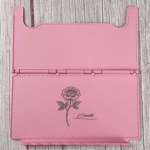 May include: A light pink foldable stand featuring a hinged design with scalloped edges. The lower panel displays a dark grey line art illustration of a single rose with a stem and leaves, accompanied by the name Cindi written in a cursive script.