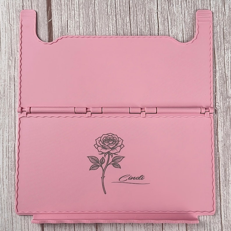 May include: A light pink foldable stand featuring a hinged design with scalloped edges. The lower panel displays a dark grey line art illustration of a single rose with a stem and leaves, accompanied by the name Cindi written in a cursive script.