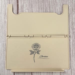 May include: A beige, rectangular, foldable plastic stand featuring a hinged design. The front panel displays a dark grey line art illustration of a single rose with a stem and leaves, accompanied by the name Christine written in a cursive script.