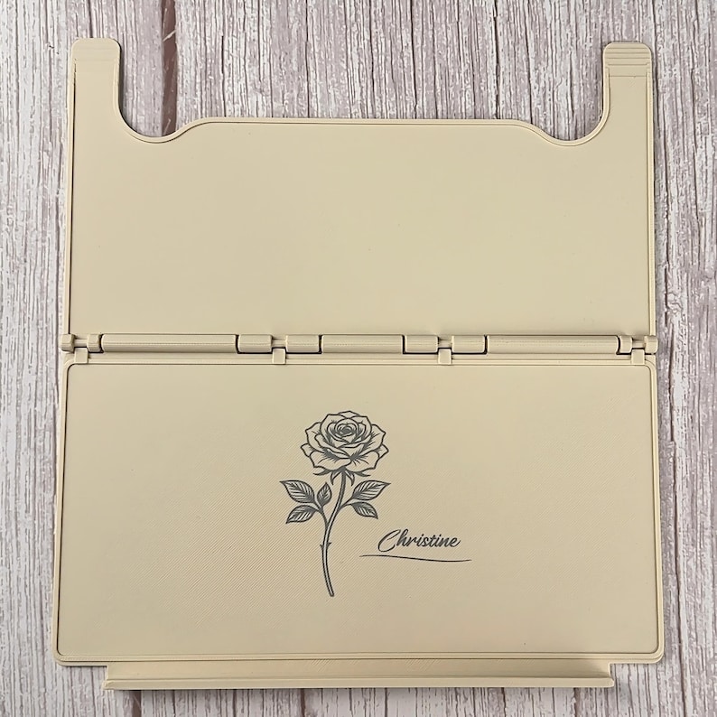 May include: A beige, rectangular, foldable plastic stand featuring a hinged design. The front panel displays a dark grey line art illustration of a single rose with a stem and leaves, accompanied by the name Christine written in a cursive script.