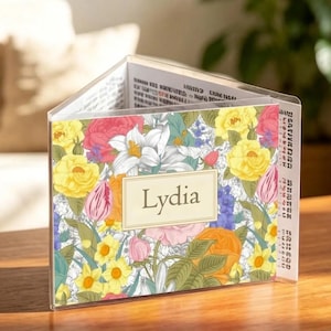 Peut inclure: Un présentoir en forme de livre en acrylique transparent avec un motif floral composé de fleurs jaunes, roses et rouges. Le nom "Lydia" est affiché sur une étiquette rectangulaire crème. Le livre est ouvert, révélant du texte à l'intérieur.