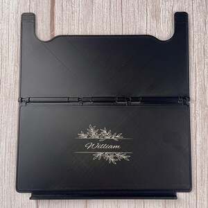 May include: A black foldable book stand featuring a central hinge and a rectangular base. The front panel is engraved with the name William in a cursive script, flanked by decorative botanical leaf illustrations above and below the text.