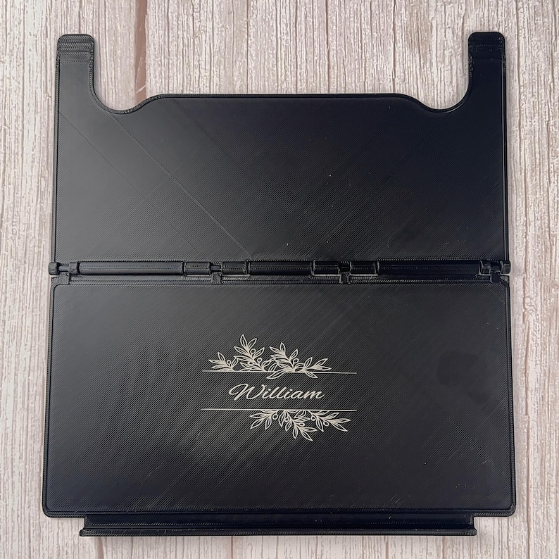 May include: A black foldable book stand featuring a central hinge and a rectangular base. The front panel is engraved with the name William in a cursive script, flanked by decorative botanical leaf illustrations above and below the text.