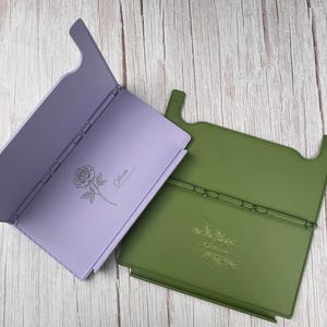 May include: Two rectangular folding stands, one lavender and one olive green. The lavender stand features a rose illustration and the name Olivia. The olive green stand features a botanical branch illustration and the name Kathleen engraved on them.