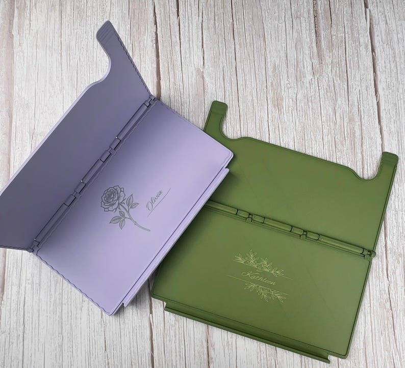 May include: Two rectangular folding stands, one lavender and one olive green. The lavender stand features a rose illustration and the name Olivia. The olive green stand features a botanical branch illustration and the name Kathleen engraved on them.