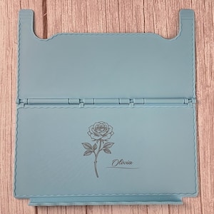 May include: A light blue foldable stand featuring a hinged design with a scalloped edge border. The lower panel displays a dark grey engraved illustration of a single rose with a stem and leaves, accompanied by the name Olivia in script text.