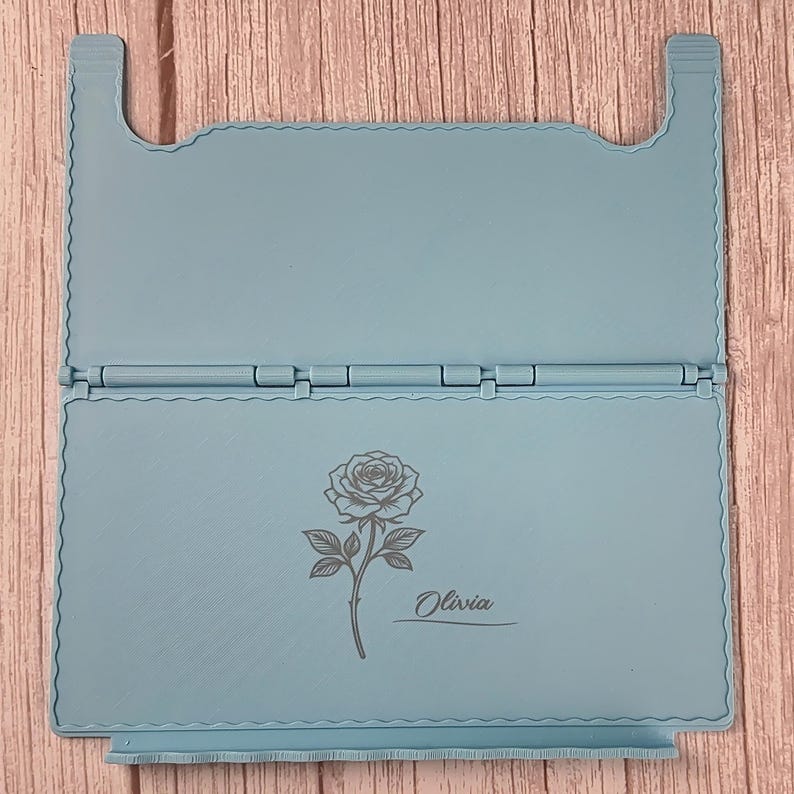 May include: A light blue foldable stand featuring a hinged design with a scalloped edge border. The lower panel displays a dark grey engraved illustration of a single rose with a stem and leaves, accompanied by the name Olivia in script text.