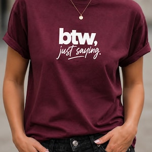 Puede incluir: Una camiseta de color granate de manga corta con el texto impreso en blanco "btw, just saying.". La prenda tiene cuello redondo y mangas remangadas, combinada con vaqueros negros. Se observa un collar fino dorado con un pequeño colgante.