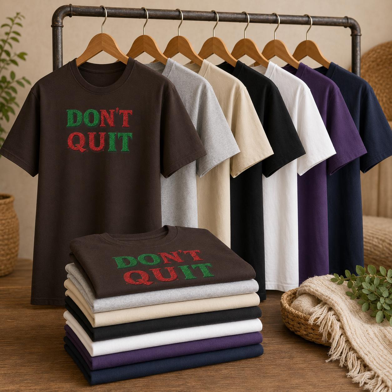 Mehrere T-Shirts in verschiedenen Farben mit ‚DON’T QUIT‘ Aufdruck auf Holzbügeln und gefaltet auf Holztisch