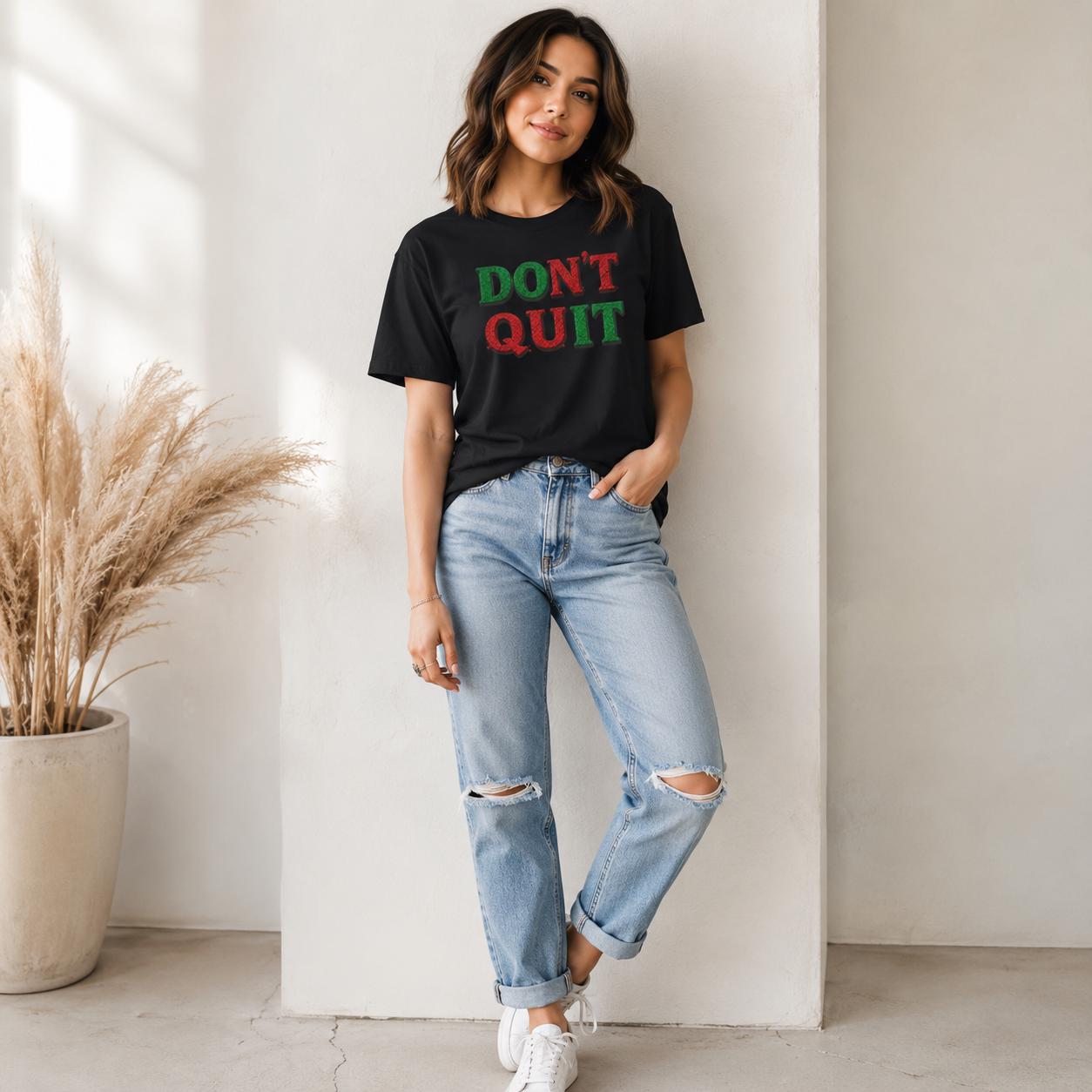 Frau trägt schwarzes ‚DON’T QUIT‘ T-Shirt kombiniert mit blauen Jeans vor weißer Wand