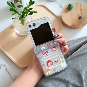 May include: A clear, light blue phone case with a flip design, adorned with sushi, bear, and strawberry graphics. The case features a house-shaped design with a blue and white checkered pattern and red bows. The phone screen is black.