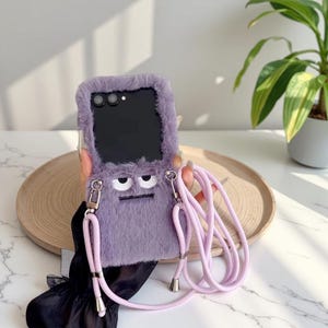 Può includere: Una custodia per telefono color lavanda con una texture pelosa e un design a forma di cartone animato. La custodia ha un'area dello schermo nera e un cinturino lavanda abbinato. La custodia è progettata per proteggere il telefono aggiungendo un'estetica unica.