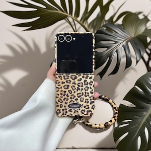 Puede incluir: Una funda de teléfono con estampado de leopardo en beige y marrón con una correa de muñeca a juego. La funda tiene un anillo de metal dorado para la correa. La pantalla del teléfono es negra.