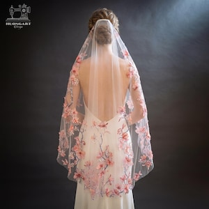 May include: A bridal veil with pink floral embellishments and a matching floral-patterned wedding dress. The veil is sheer and drapes over the shoulders, adorned with 3D flowers and sparkling accents. The dress features a low back and delicate floral embroidery.