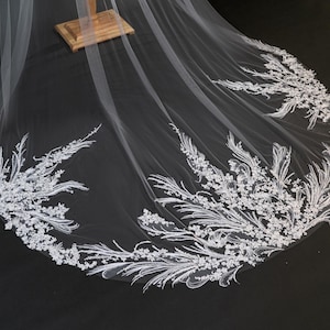 Può includere: Un velo da sposa bianco con intricati dettagli in pizzo floreale e a foglie lungo il bordo. Il velo è drappeggiato su un supporto in legno, mettendo in mostra il design e la consistenza delicati. Il design del velo è visibile su uno sfondo scuro.