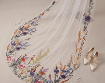 May include: A white wedding veil with colorful floral embroidery along the edges. The embroidery features blue, orange, yellow, and purple flowers and greenery. Two pairs of white satin heels and a few decorative pins are also in the image.