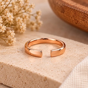 Puede incluir: Un anillo abierto de tono oro rosa pulido que presenta una banda lisa y redondeada con una abertura en la parte frontal. La superficie metálica tiene un acabado reflectante, mostrando un diseño simple y continuo de forma circular.