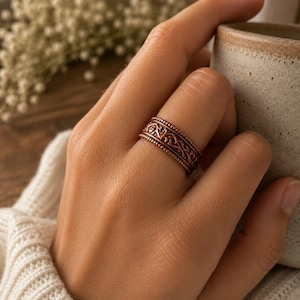 Puede incluir: Un anillo de metal en tono cobre que presenta un intrincado patrón de volutas en relieve en la banda central, bordeado por dos filas delgadas de detalles de cuentas elevadas a lo largo de los bordes superior e inferior.