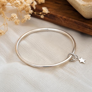 Peut inclure: Un bracelet jonc de couleur argentée polie doté d'un anneau circulaire lisse. Une petite breloque en forme d'étoile à cinq branches est fixée au cerceau principal par deux anneaux de jonction interconnectés, formant un accessoire de poignet.